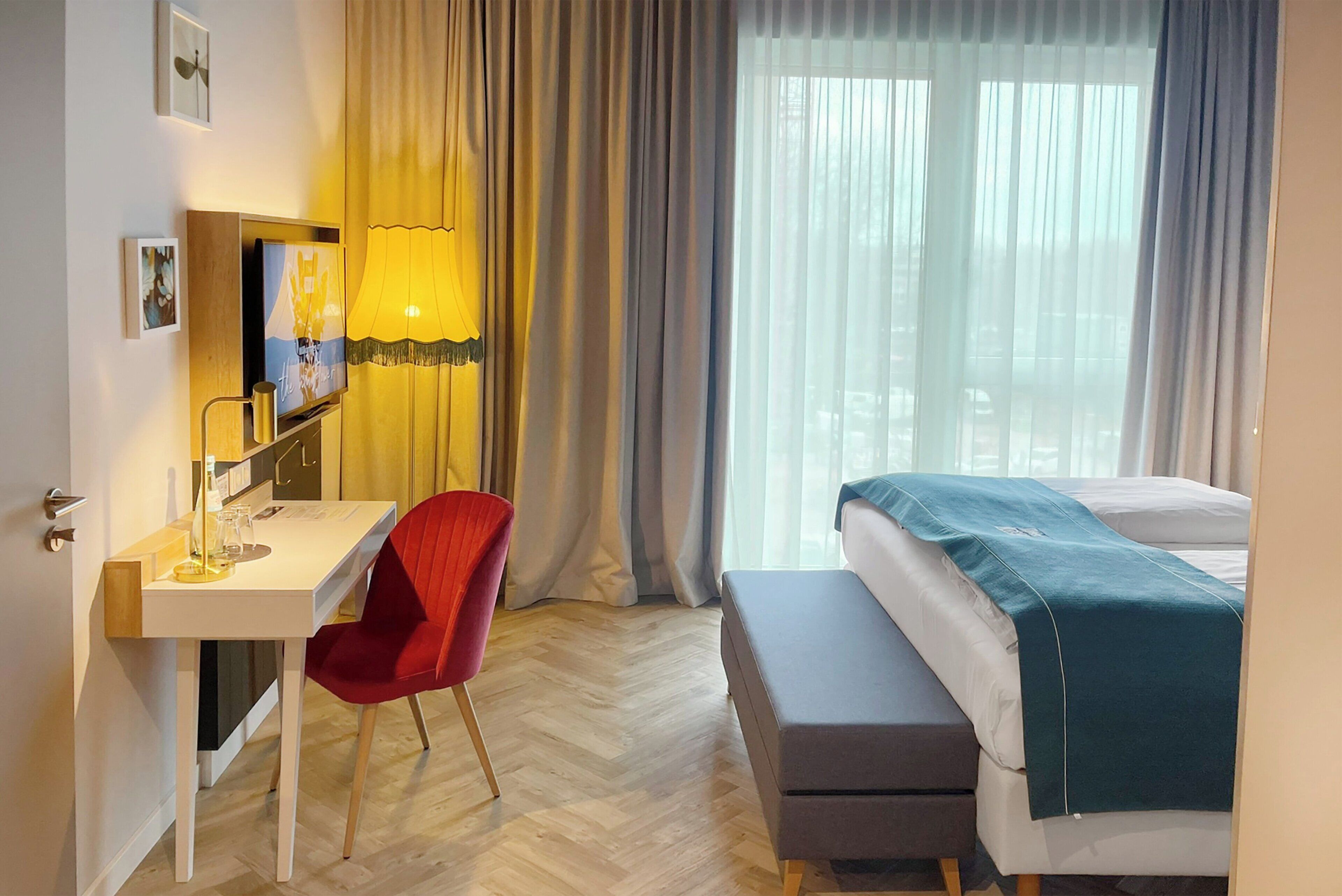 Habitación de hotel con cama, escritorio, silla, lámpara y ventana con cortinas.
