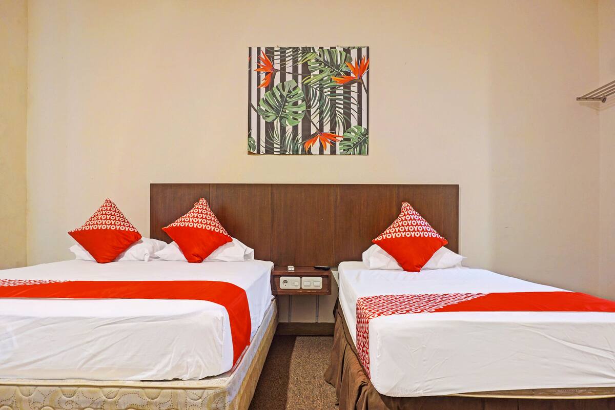 Ein Hotelzimmer mit zwei Betten, jeweils mit weißer Bettwäsche und roten Kissen, einem hölzernen Kopfstück, einem Nachttisch mit Telefon und Fernbedienung sowie einem Bild von tropischer Vegetation an der Wand.