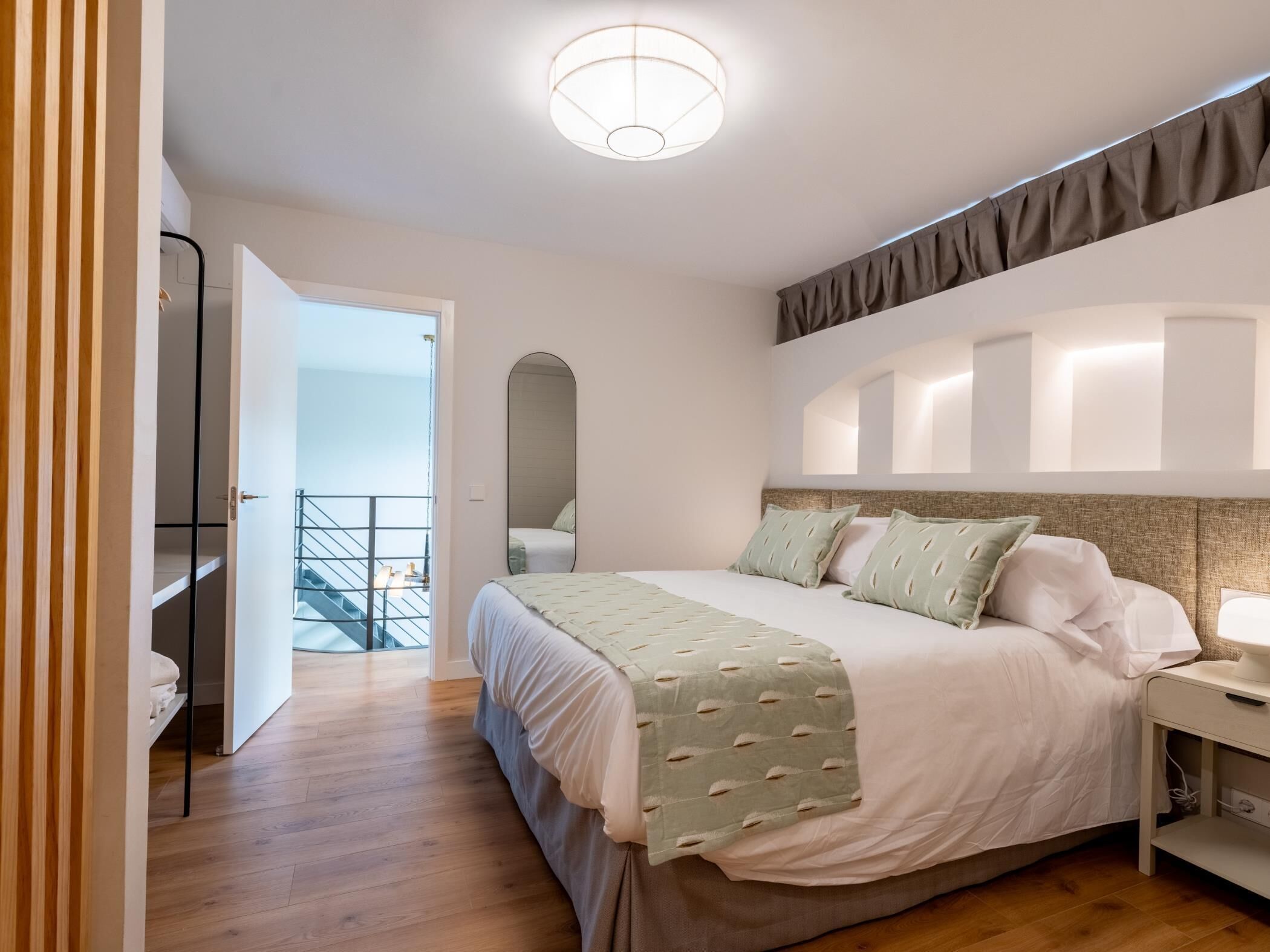 Quarto moderno com uma cama grande, mesa de cabeceira, espelho e vista para uma varanda.