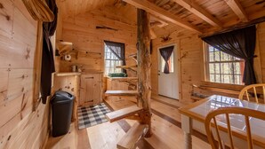 A wooden cabin interior with a dining table, chairs, a kitchen area, and a large window. - Dogwood Cabin - Off Grid (Chesterhill)