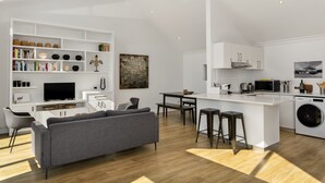 A modern kitchen with white cabinetry, a central island with bar stools, a built-in washing machine, and a dining area with a gray sofa and a flat-screen TV. - Burnside 302 (Cape Town)