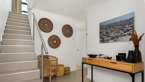 A staircase with a metal railing, a wicker chair, a wooden table with decorative items, and a cityscape painting on the wall. - Burnside 302 (Cape Town)