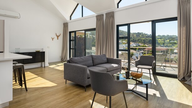 A modern living room with a grey sofa, a glass coffee table, and a dining area with a white table and chairs. Large windows offer a view of the outdoors. - Burnside 302 (Cape Town)