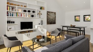 A modern living room with a sofa, coffee table, and a TV on a white cabinet. There are books, decorative items, and a painting on the shelves. - Burnside 302 (Cape Town)
