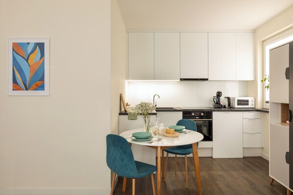 Una cocina compacta con armarios blancos, microondas, cafetera y comedor con sillas color turquesa y una mesa blanca.