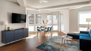 A modern living room with a dining area, a flat-screen TV mounted on the wall, and large windows providing natural light. - Trevi Apartments Tychy (Tychy)