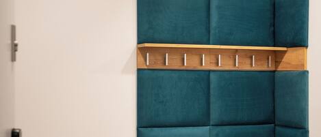 Une pièce d’angle avec un banc recouvert d’un tissu turquoise, une étagère en bois avec des crochets et un radiateur.