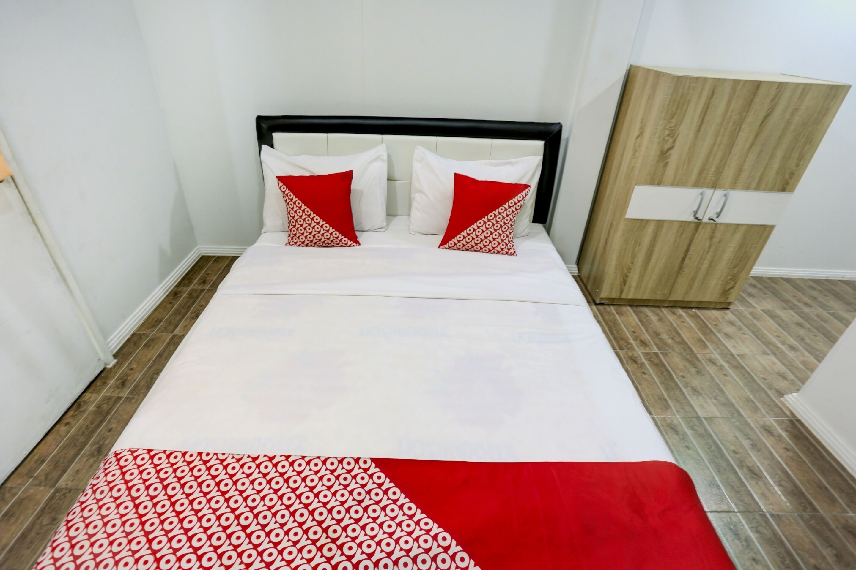 A neatly made bed with a white and red patterned bedspread and pillows, a wooden wardrobe, and a tiled floor.