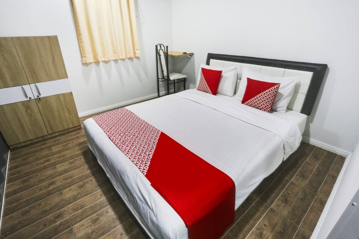 Un lit bien fait, recouvert d’une couverture blanche et rouge, une armoire en bois et une chaise avec une petite table placée à côté.