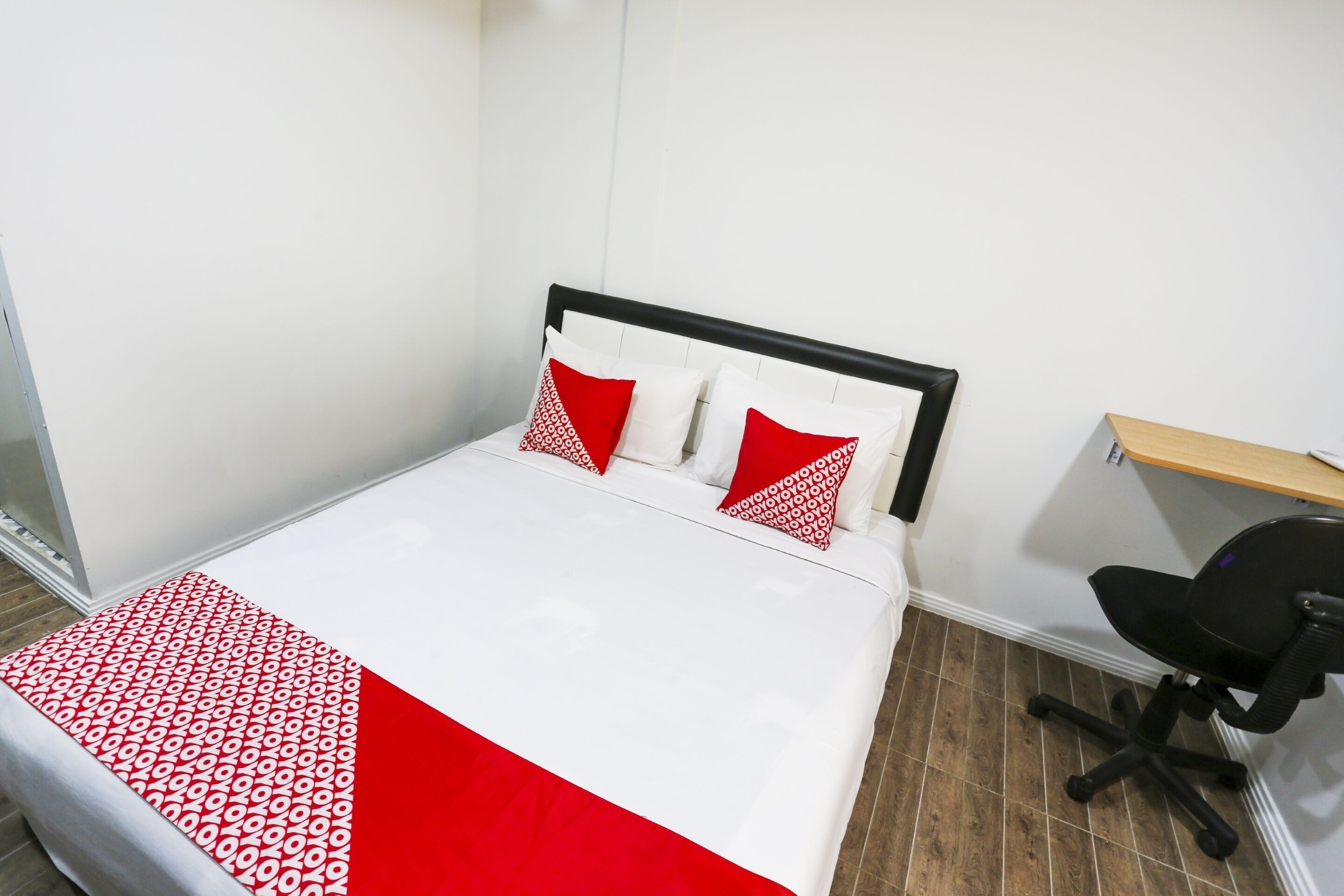 A neatly made bed with a white and red bedspread, two red pillows, and a black office chair in a room with white walls and wooden flooring.