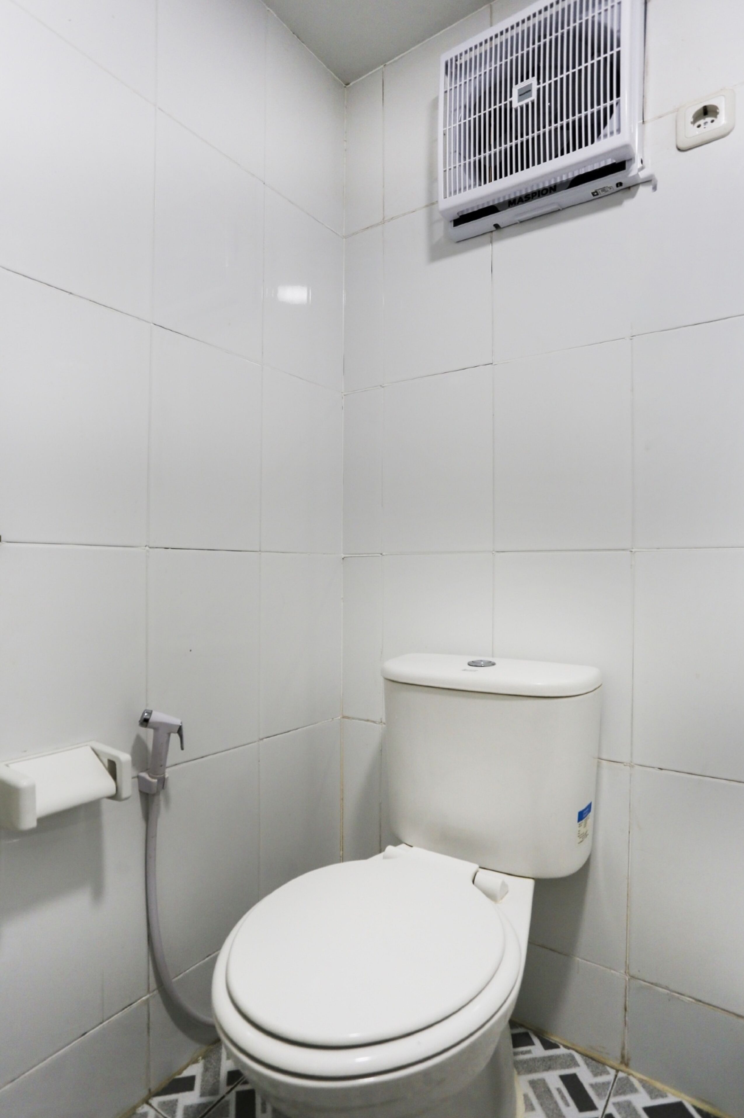 Een kleine, witte betegelde badkamer met toilet, toiletrolhouder en een witte airconditioner die aan de muur is bevestigd.