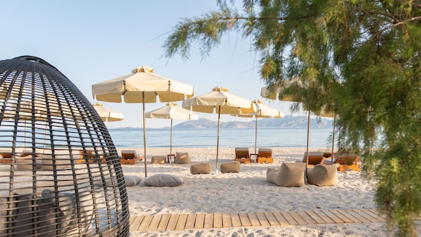 Una playa con sillas y sombrillas de playa, una terraza de madera y vistas al mar y a las montañas.