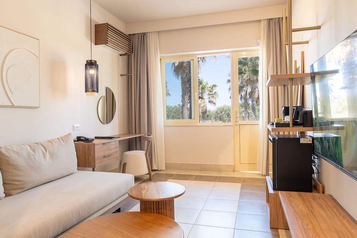 Ein modernes Hotelzimmer mit einer Couch, einem kleinen runden Holztisch, einem Schreibtisch mit Stuhl, einer Minibar und einem großen Fenster mit Blick auf Palmen.