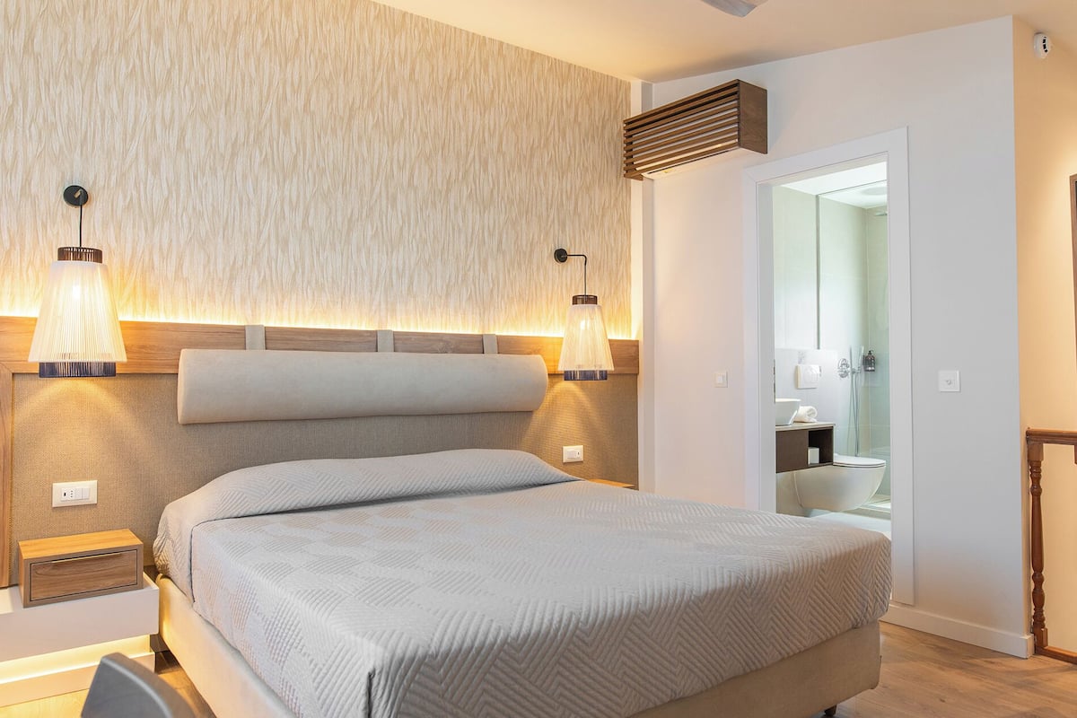 Ein modernes Schlafzimmer mit einem großen Bett, zwei Wandleuchten, einem Holz-Kopfteil und einer sichtbaren Badezimmertür.