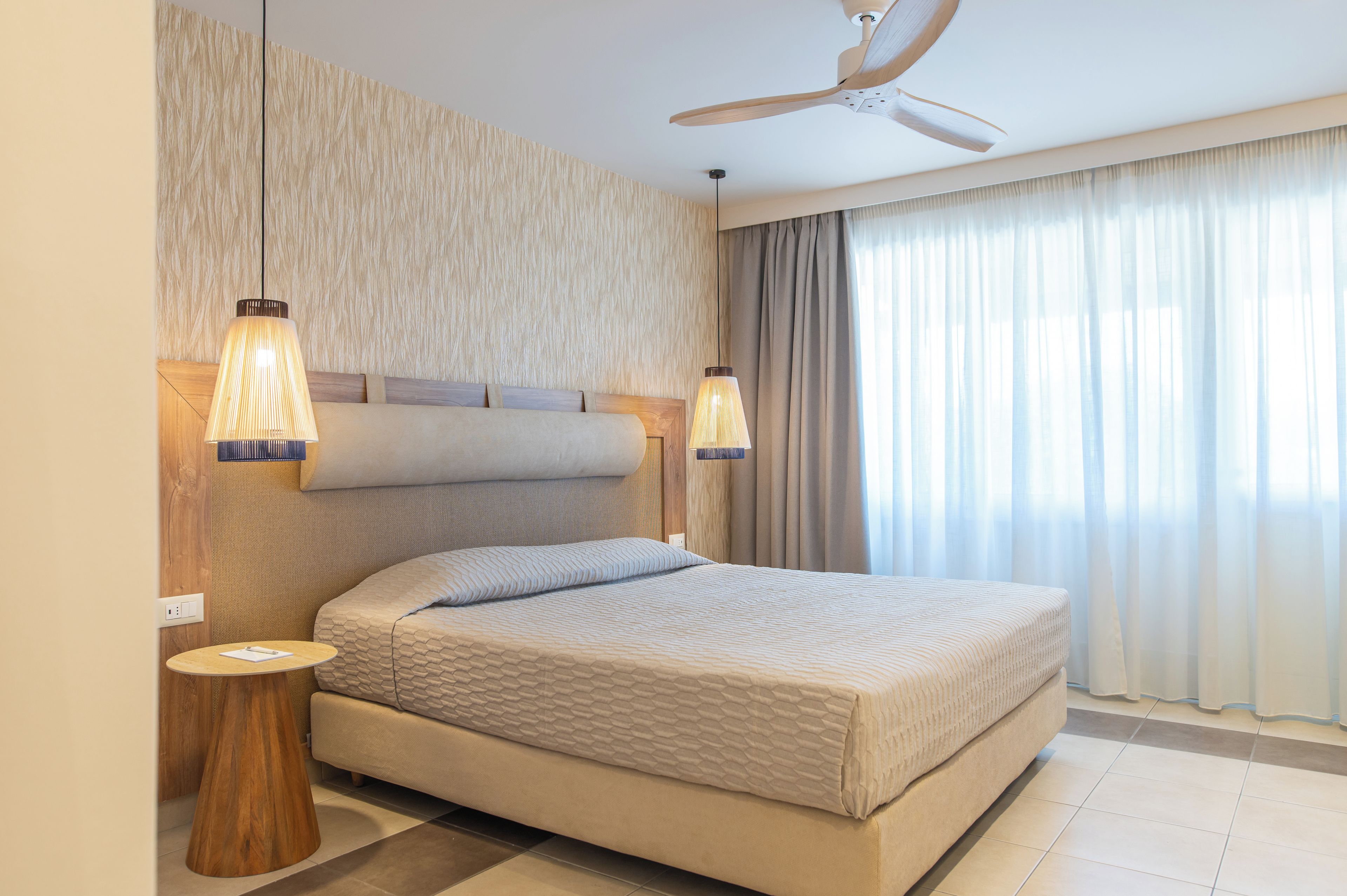 A hotel room with a bed, two pendant lights, a ceiling fan, and a large window with curtains.