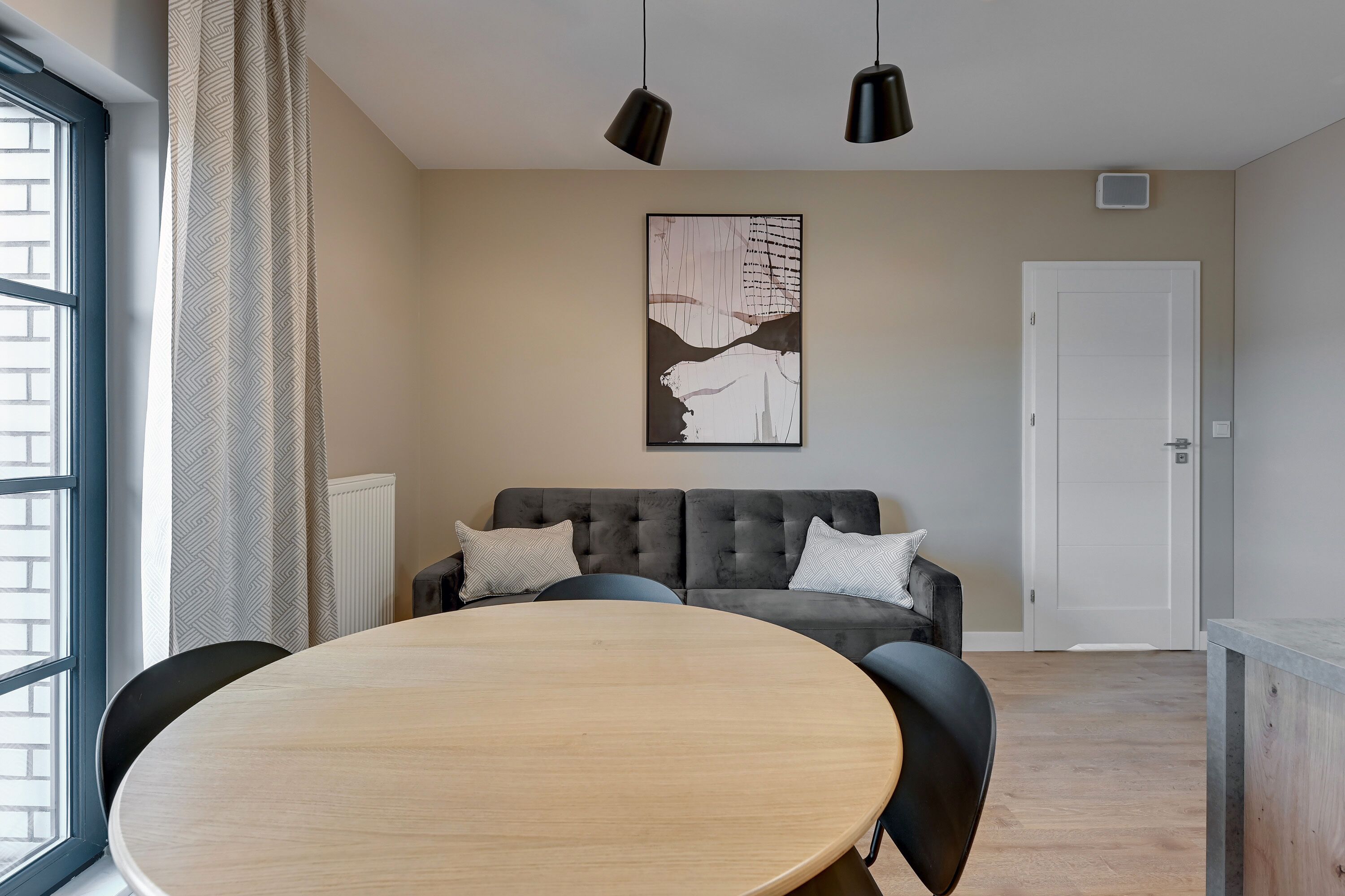 A modern living room with a round wooden table, a grey sofa with two pillows, and a large abstract painting on the wall.