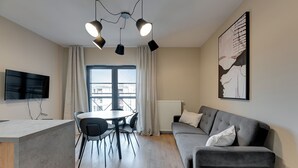 Een moderne woonkamer met een grijze bank, een eettafel met stoelen, een flatscreen televisie en een groot raam met gordijnen.