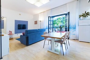 A modern living room with a blue sofa, a wooden dining table with chairs, a flat-screen TV mounted on a wall, and a large window with a view of greenery.