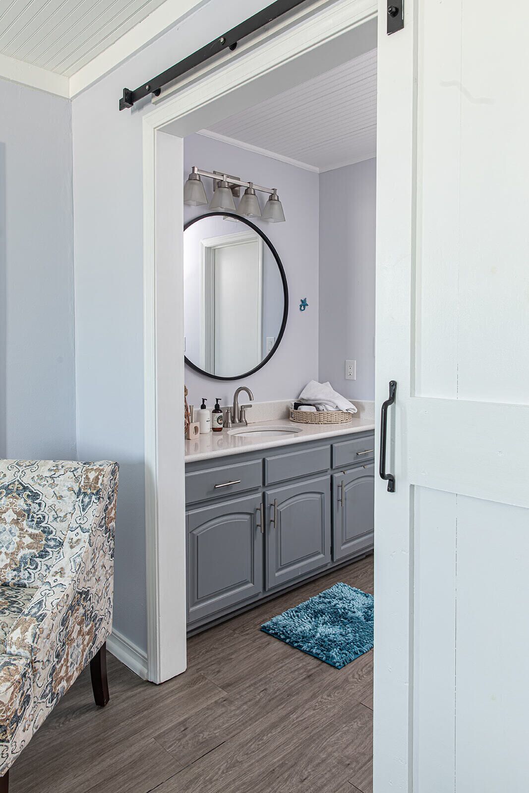 A bathroom with a large mirror, grey cabinets, a white sink, and a blue rug.