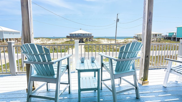 A wooden deck with two chairs and a table, overlooking a beach with a house on stilts.