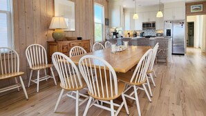 Un vaste espace de salle à manger avec une table en bois et des chaises blanches, une cuisine équipée d’appareils électroménagers modernes et une grande fenêtre laissant entrer la lumière naturelle.