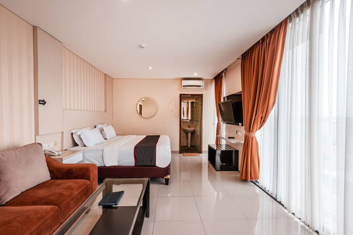 Habitació d'hotel moderna amb llit, sofà, televisió i bany visible per una porta oberta.