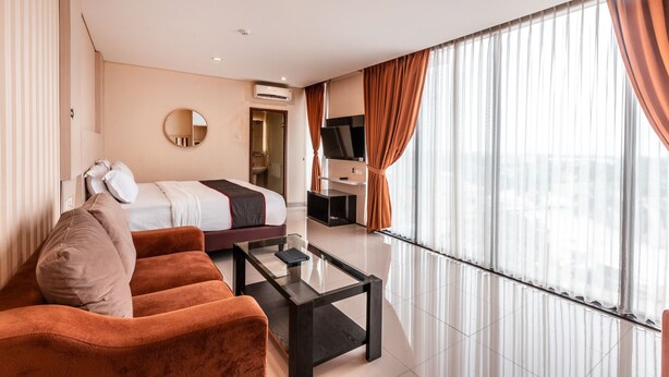 A modern hotel room with a bed, a sofa, a coffee table, a TV, and large windows with curtains.