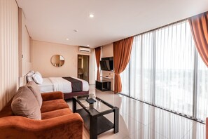 A modern hotel room with a bed, a sofa, a coffee table, a TV, and large windows with curtains. - Collection O Hotel Igloo (Cikarang)