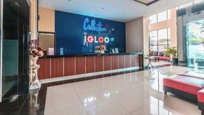 A hotel reception area with a modern design, featuring a reception desk, a seating area with a red sofa, and a large window providing a view of the outdoors.