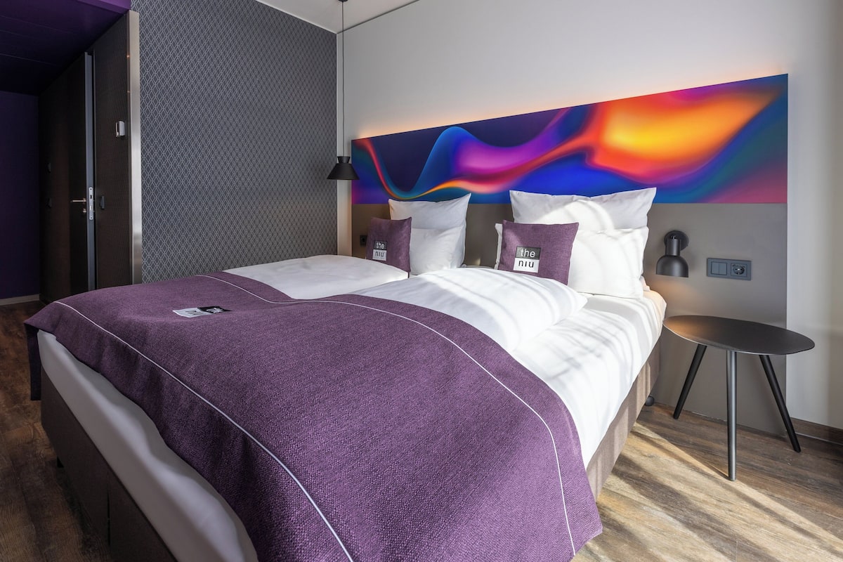 A modern hotel room with a large bed, a vibrant abstract wall art, and a small side table.