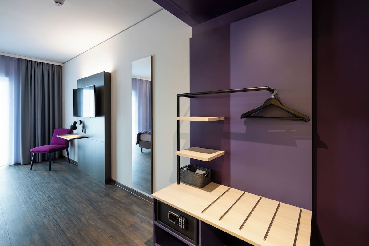 A modern hotel room with a purple accent wall, a wooden desk with a hanging light, a chair, and a mirror.