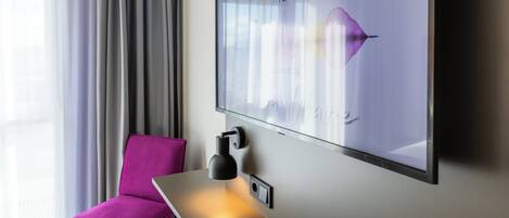 A modern hotel room with a large window, a purple chair, a desk with a lamp, and a wall-mounted TV displaying a graphic.