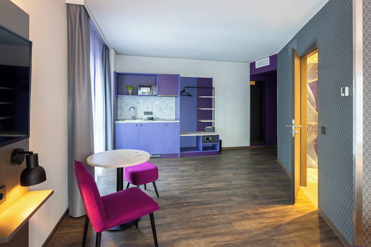 A modern hotel room with a kitchen area, a dining table with chairs, and a walk-in closet.