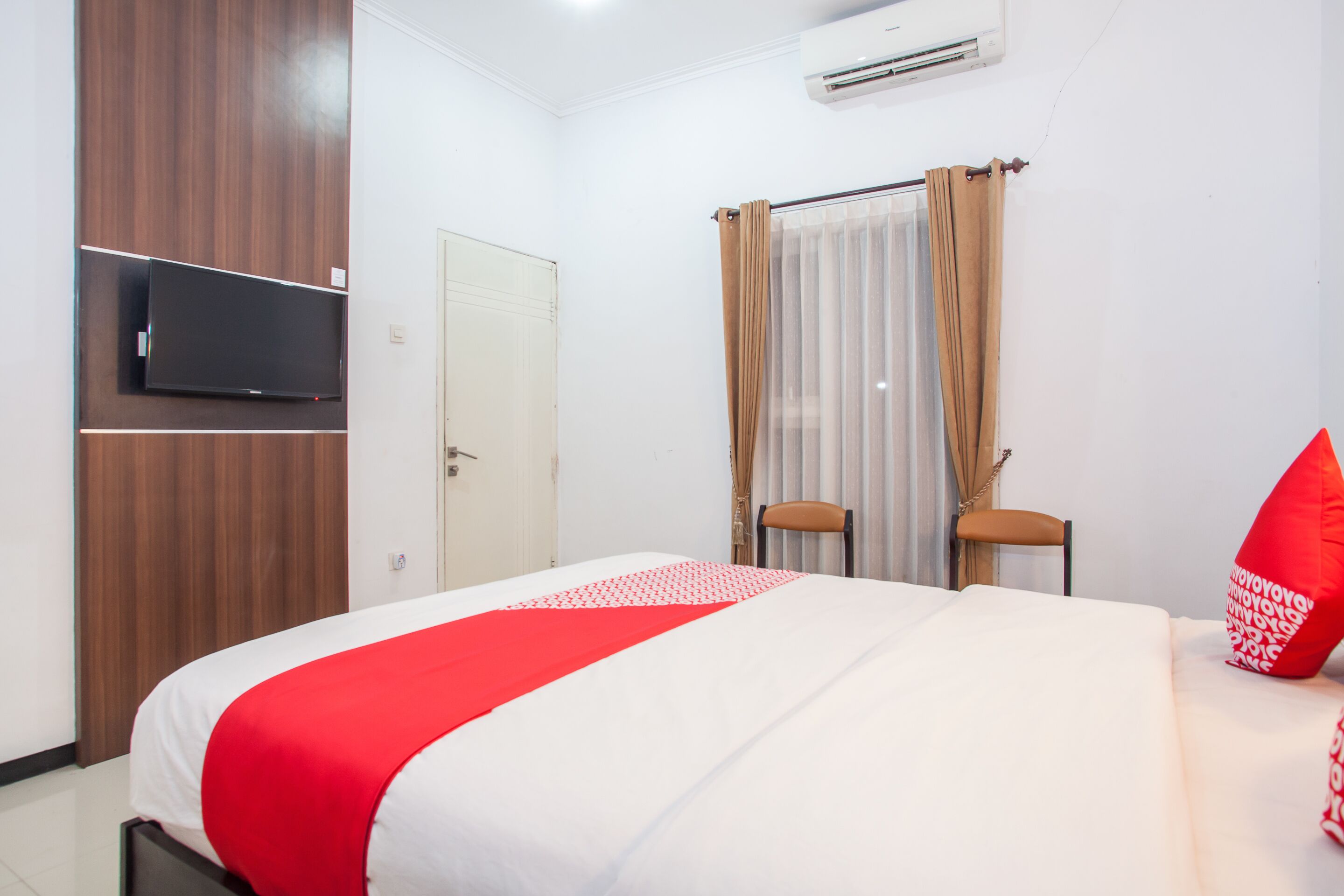 Una camera d'albergo con un letto, una televisione a muro, una porta e una finestra con tende.