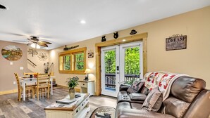 Una sala de estar acogedora con un sofá de cuero, una mesa de comedor con sillas, una zona de cocina y un amplio ventanal con vistas a la vegetación.