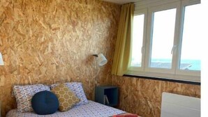 A bedroom with a wooden wall, a bed with patterned bedding, a bedside lamp, and a window with curtains. - Apartment for 2 people - Ocean view😍 (Woignarue)