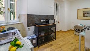 Une petite cuisine équipée d’un évier, d’un four à micro-ondes et d’un petit placard. On y trouve une fenêtre avec des rideaux, un plancher en bois et un lit recouvert d’une courtepointe à motifs.