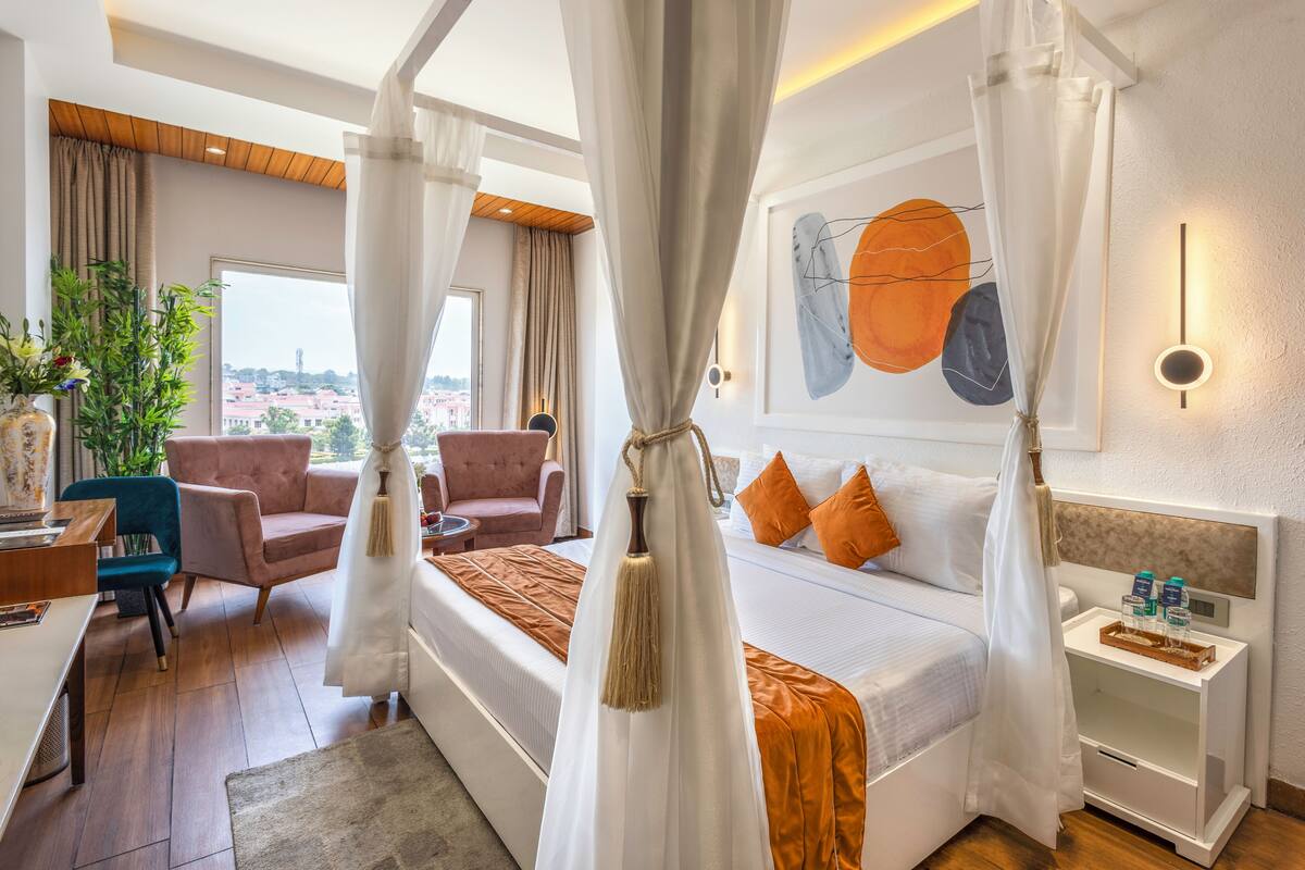 Une chambre d’hôtel avec un grand lit, un coin salon comprenant une chaise et une table, une table de chevet avec une lampe, et une vue sur la ville par la fenêtre.