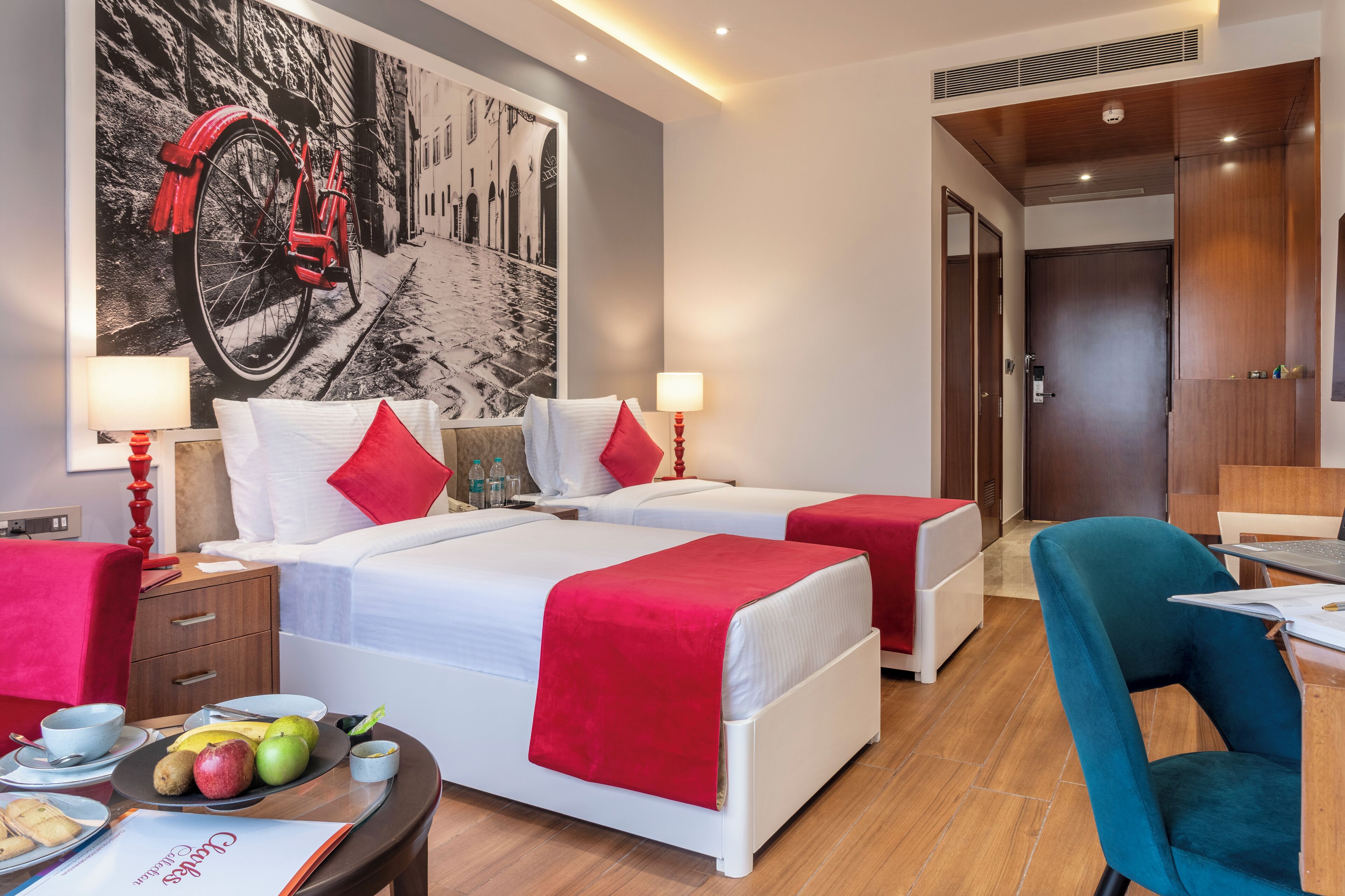 A hotel room with two beds, a dining table with breakfast, a red bicycle wall art, and a wooden door.
