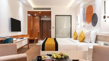 A modern hotel room with a bed, a desk, a television, and a dining area with fruit and a coffee cup.