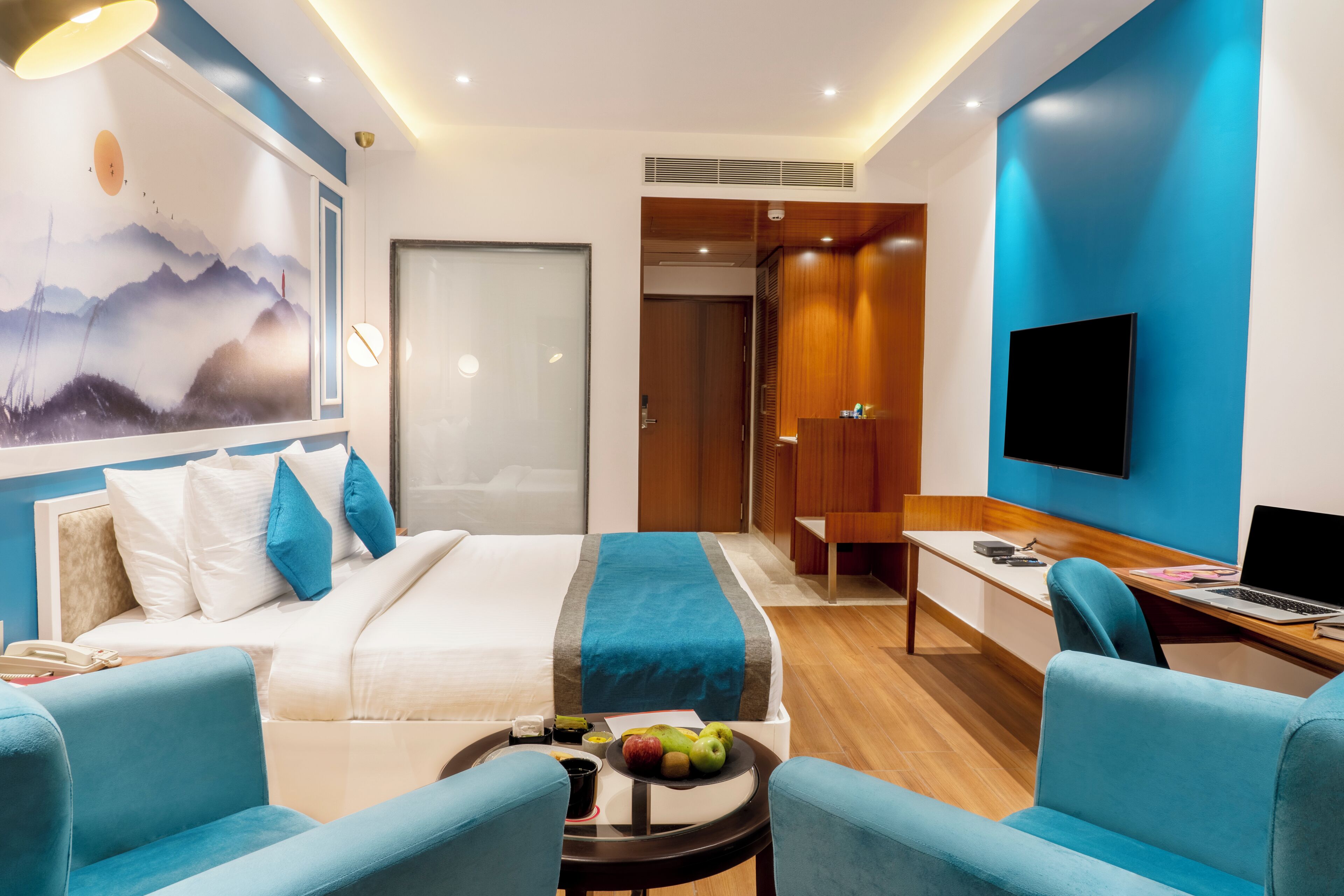 A modern hotel room with a bed, two blue armchairs, a desk with a laptop, a flat-screen TV, and a painting on the wall.