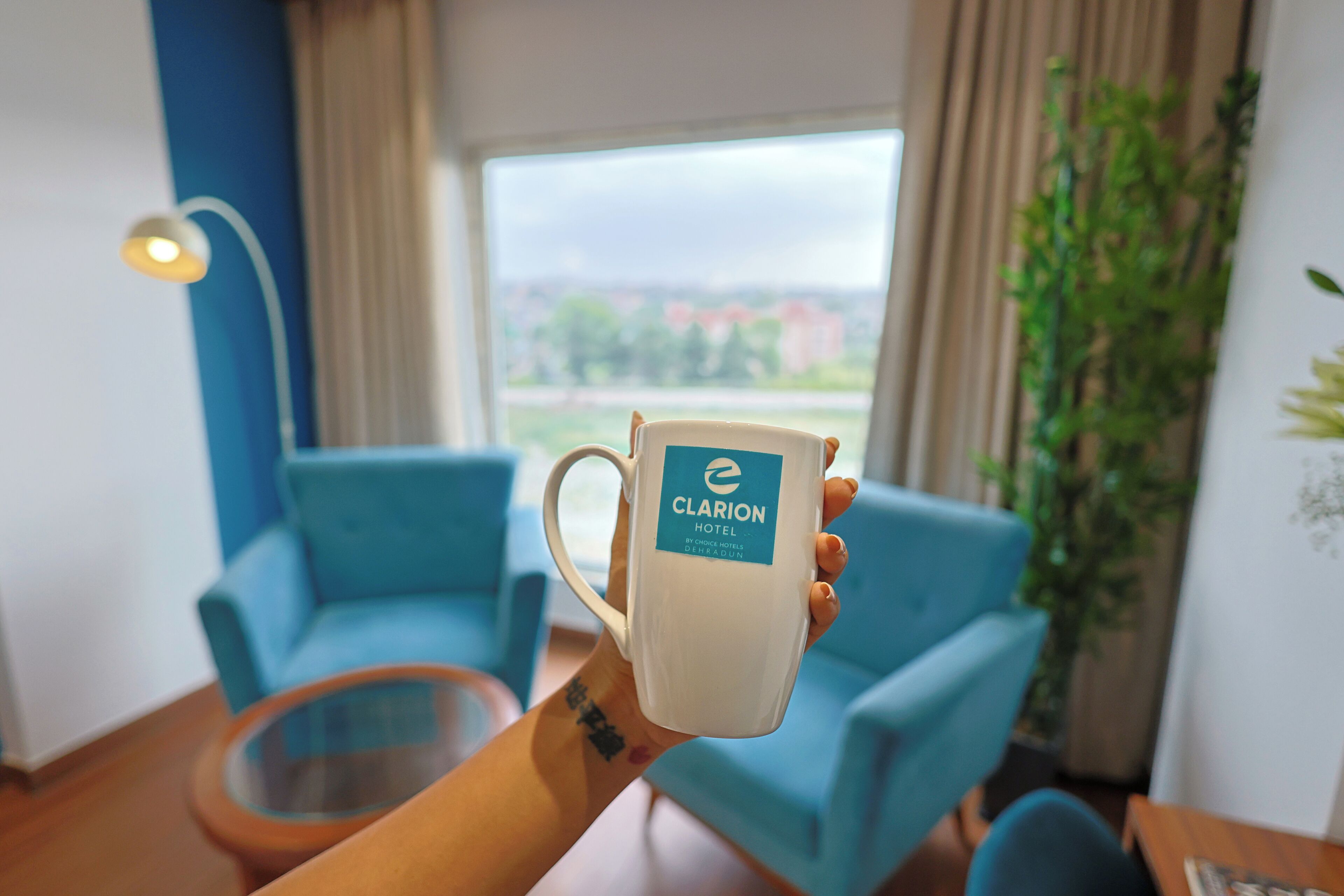 A hand holding a mug with the logo "Clarion Hotel" in a room with blue furniture and a view of buildings outside.
