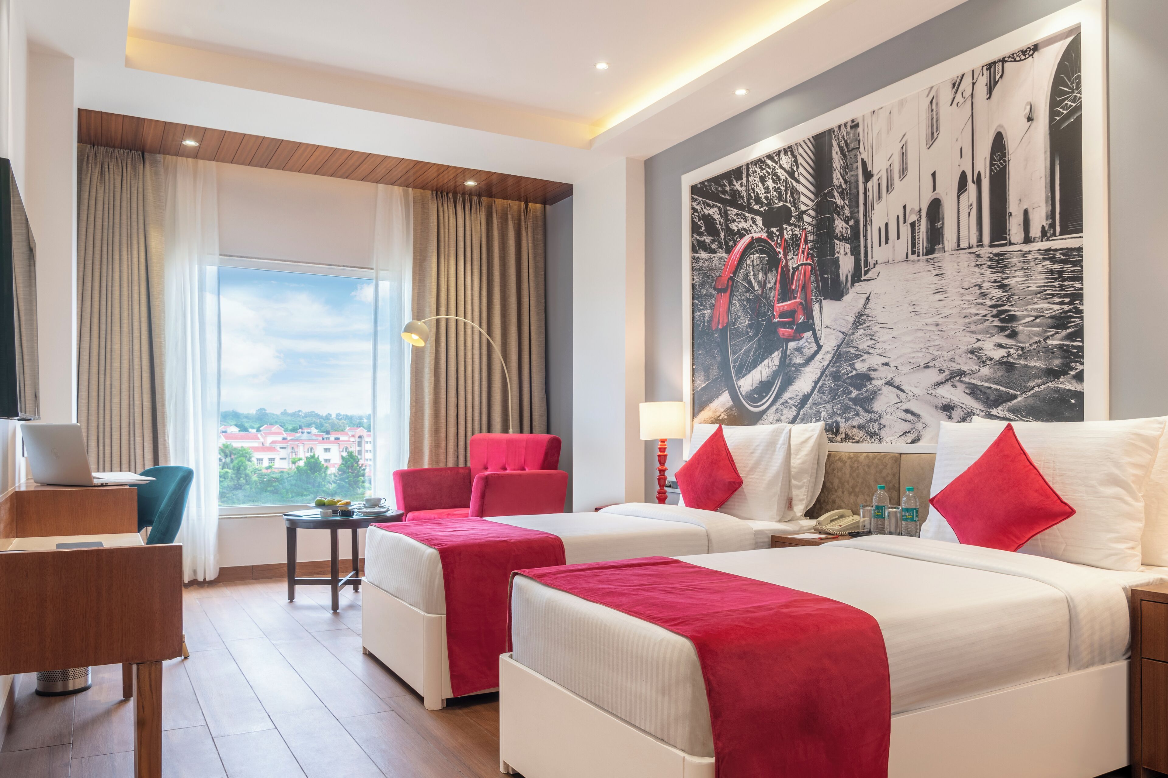 Ein Hotelzimmer mit zwei Betten, einem Schreibtisch, einem Stuhl, einem Fenster mit Vorhängen und einem großen Wandbild.