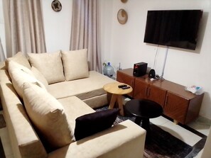 A living room with a beige sofa, a small round table, a TV on the wall, and a wooden cabinet. - 1 Bed Flat - Sleeps 2 - Parking - Wifi (Nairobi)