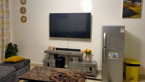Ein Wohnzimmer mit Flachbildfernseher, grauem Sofa, Couchtisch, Kühlschrank und Mülleimer.