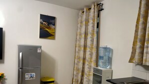 Eine kleine Küche mit Kühlschrank, Wasserspender, Mülleimer und einem gemusterten Vorhang.