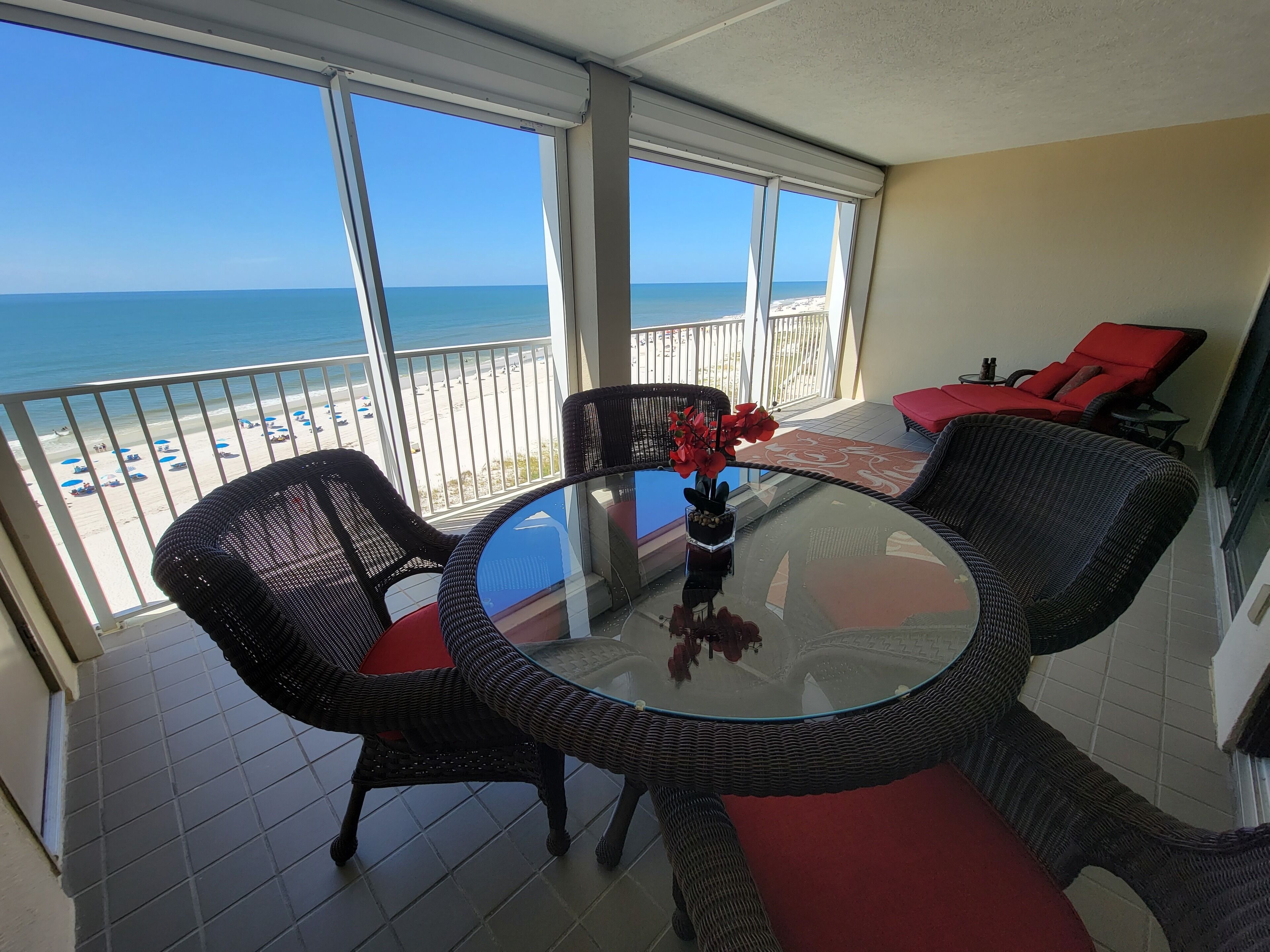 A balcony with a glass table and wicker chairs, overlooking a beach with umbrellas.