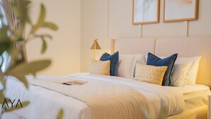 Een netjes opgemaakt bed met een witte deken en meerdere kussens, een nachtkastje met een lamp, en twee kunstwerken in lijsten aan de muur.