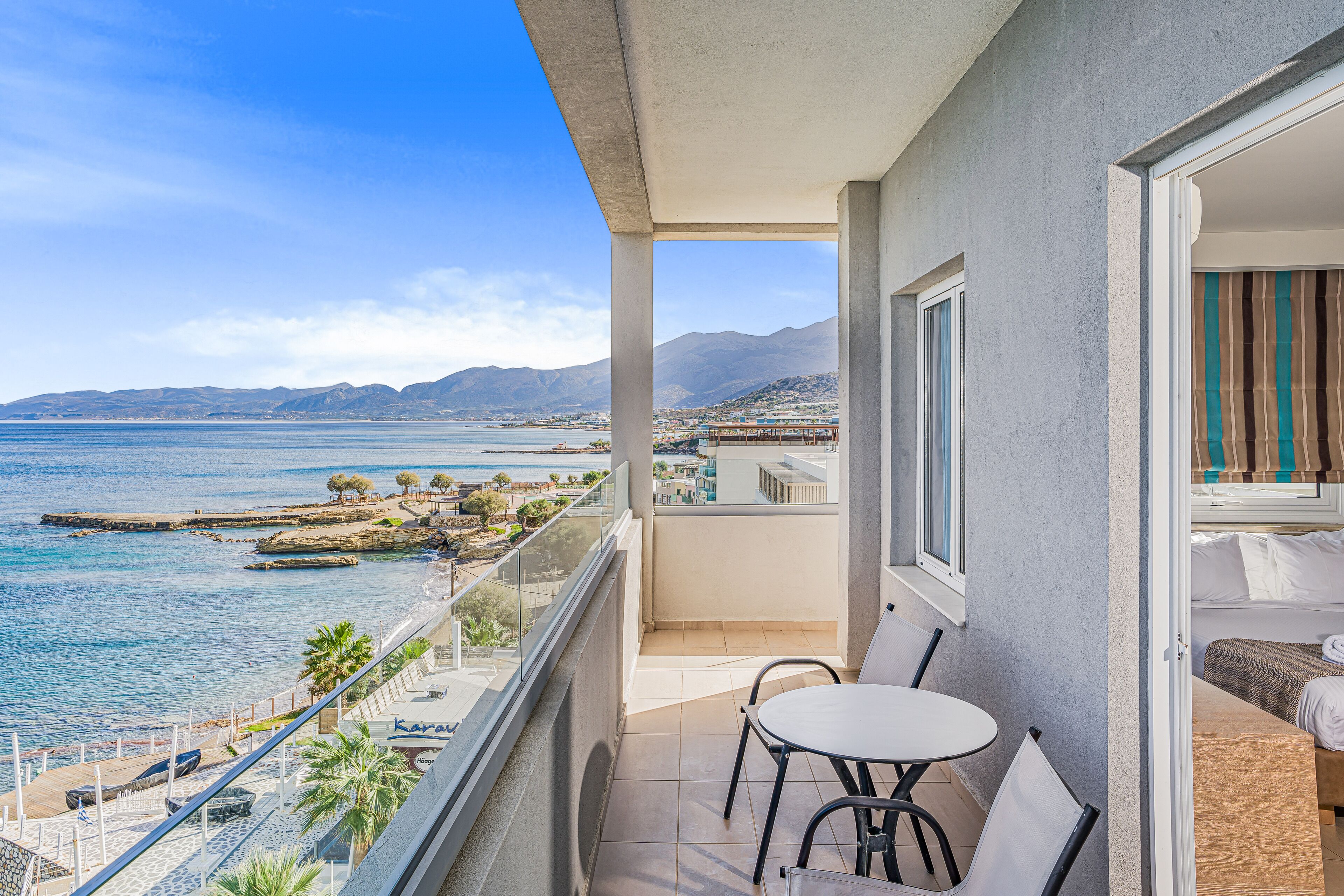 A balcony with a view of the sea and mountains, a table and chairs, and a bed with curtains.