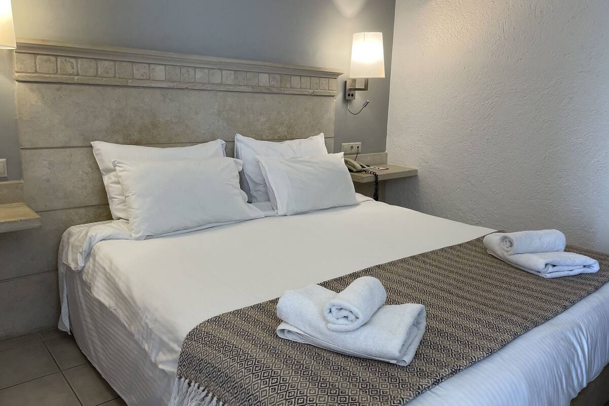 A neatly made bed with white linens and a patterned throw blanket, a bedside phone, and a wall-mounted lamp.